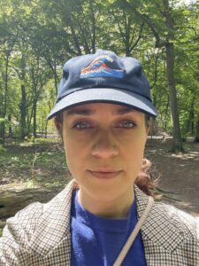 Foto von Nina Süßmilch mit blauer Cap, blauem Pullover und beig karierter Jacke im Wald.